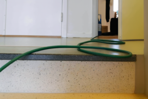 Svart slang ligger längs ett ljust plastgolv i en trappuppgång under en stamspolning i ett flerfamiljshus i Segeltorp. Golvet är nylagt och har en slät yta i beige ton. Slangen slingrar sig över ett trappsteg upp mot ett öppet rum i bakgrunden.