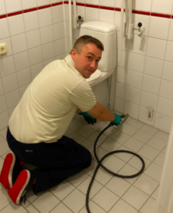 Rörmokare i badrum tittar mot kameran medan han arbetar med en spolslang vid toalett.