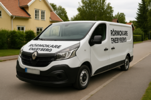 Vit Renault Trafic med texten "Rörmokare Enebyberg" på sidan, parkerad i ett villaområde i Enebyberg med blurrad registreringsskylt.